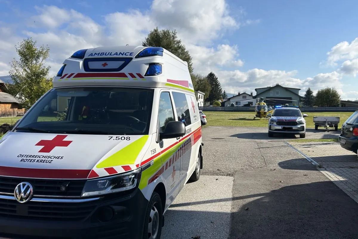 Ein Rettungswagen des Österreichischen Roten Kreuzes steht auf einem Parkplatz, daneben ein Polizeiauto und im Hintergrund ein Rettungshubschrauber auf einer Wiese bei einem sonnigen Tag.