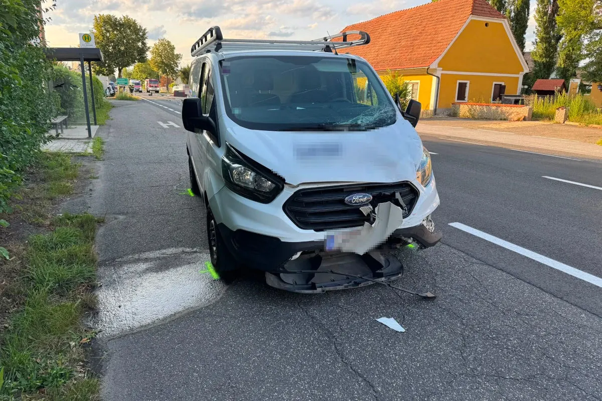 Ein weißer Ford-Transporter mit beschädigter Front steht auf einem Gehweg in einer ländlichen Umgebung neben einer Bushaltestelle.