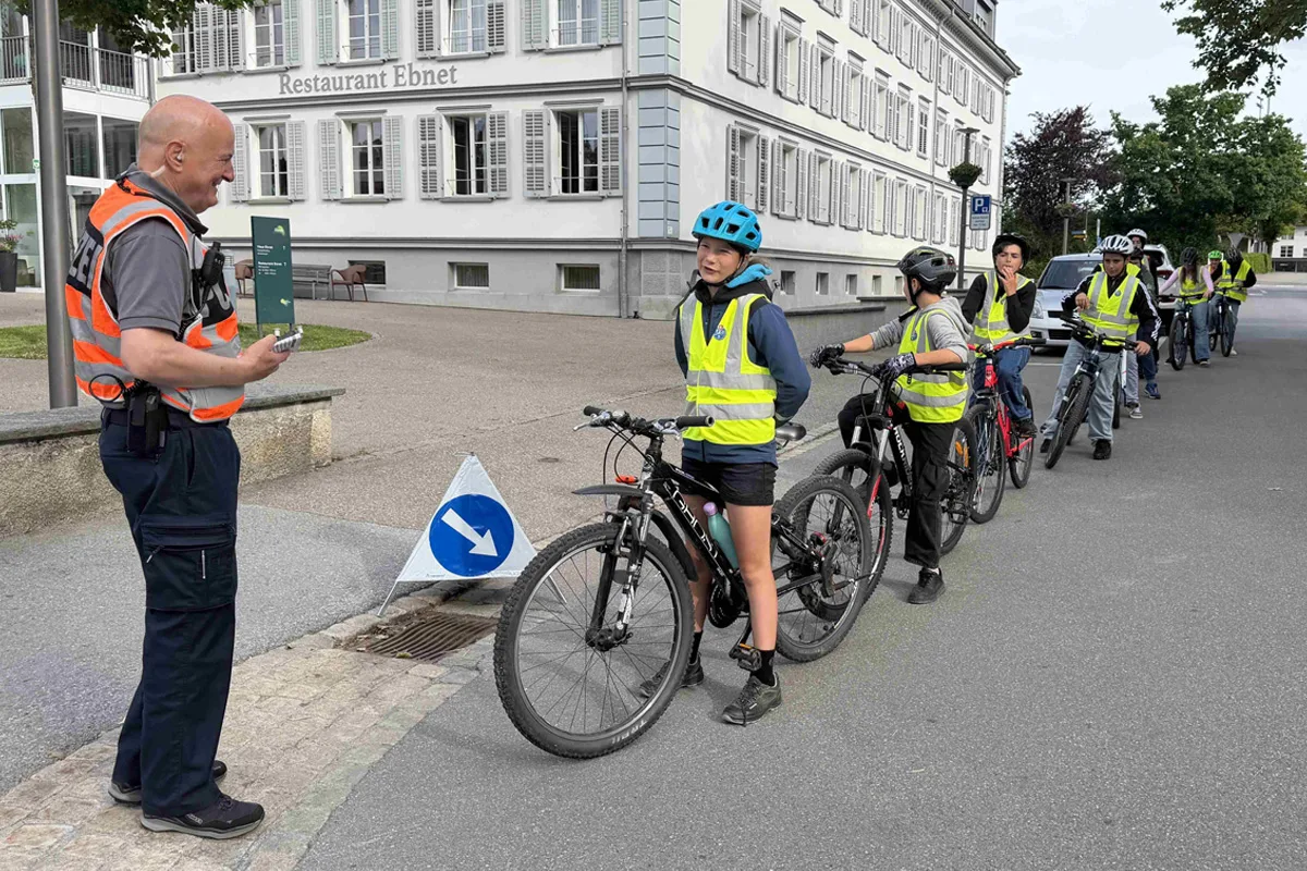 Ein Polizist in oranger Sicherheitsweste spricht mit einer Gruppe von Kindern auf Fahrrädern, die gelbe Warnwesten tragen. Die Szene spielt sich vor einem Gebäude mit dem Schild 'Restaurant Ebnet' ab.
