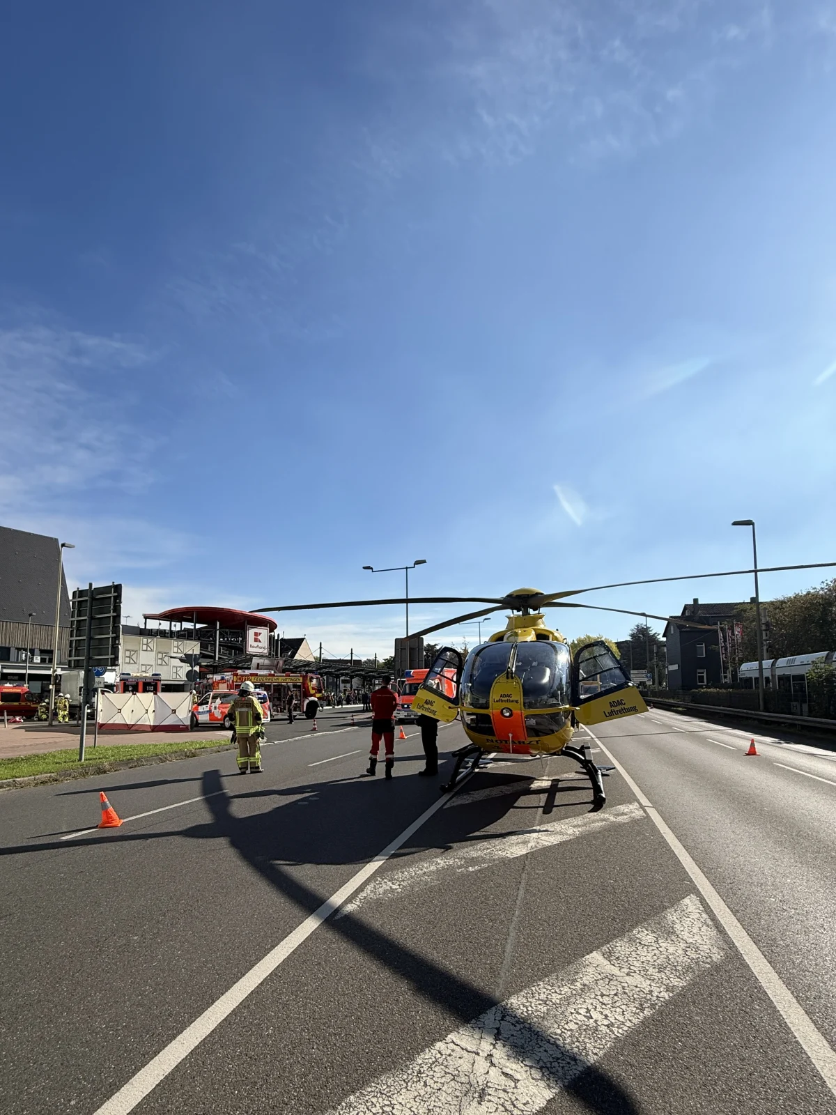 Ein gelber Rettungshubschrauber steht auf einer gesperrten Straße. Rettungskräfte und Fahrzeuge sind im Hintergrund zu sehen.