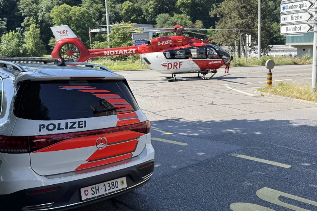 Ein Polizeifahrzeug steht nahe einem DRF-Rettungshubschrauber, der auf einer Straße gelandet ist. Im Hintergrund sind Straßenschilder und Bäume sichtbar.