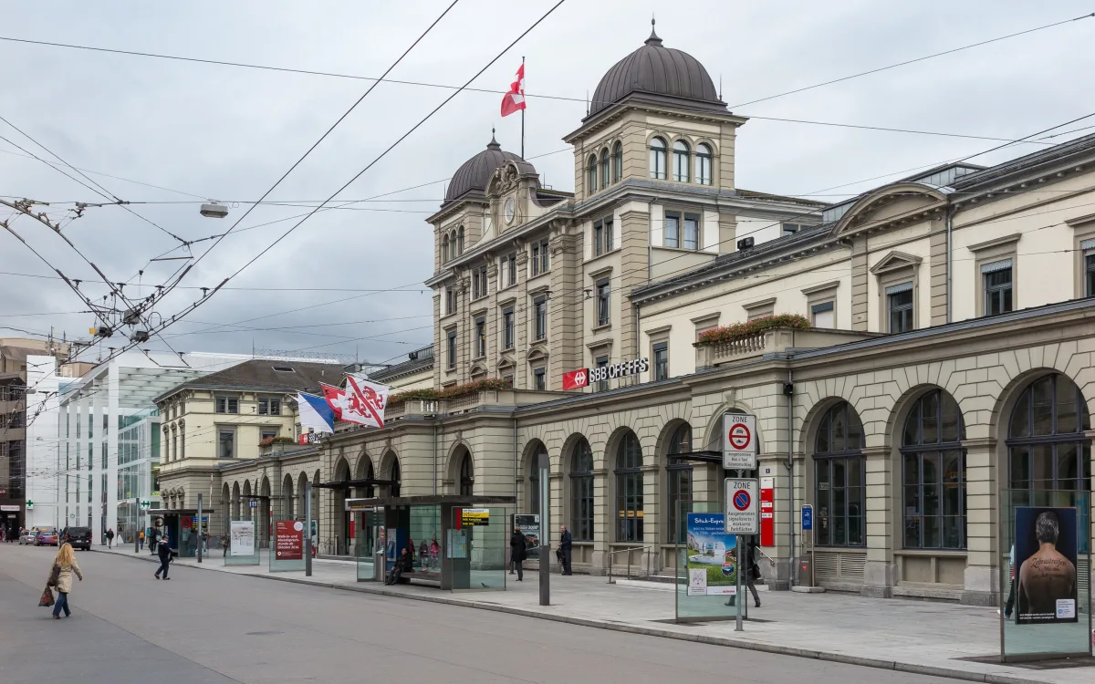 Ein belebter Hauptbahnhof mit der Schweizer Flagge auf dem Gebäude. Passanten und Straßenbahnlinien sind gut sichtbar.