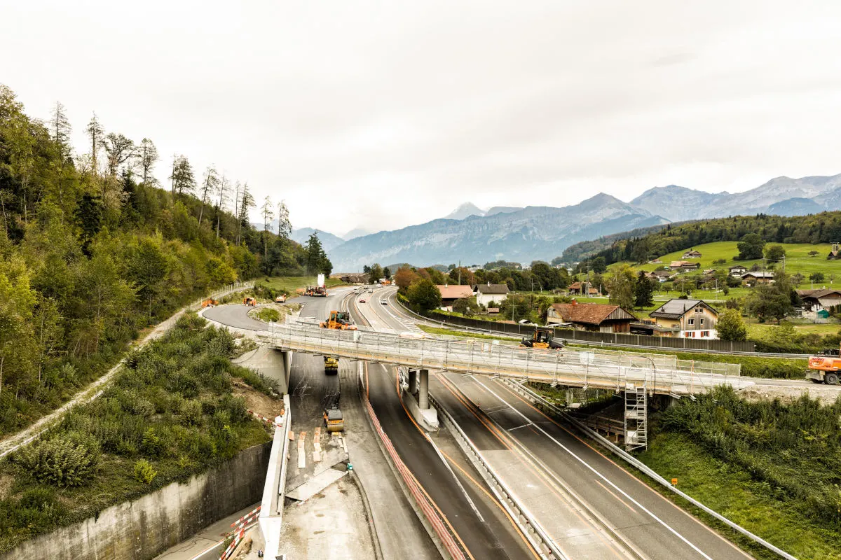 Auf einer mehrspurigen Straße finden Bauarbeiten unter einer Brücke statt. Mehrere Baustellenfahrzeuge sind in ländlicher Umgebung zu sehen.