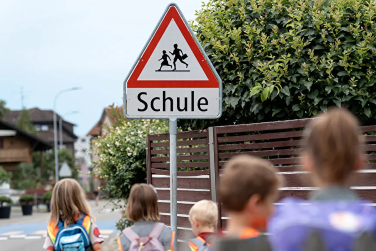 Ein Verkehrsschild mit der Aufschrift 'Schule' steht an einer Straße, während mehrere Kinder mit Rucksäcken vorbeigehen.