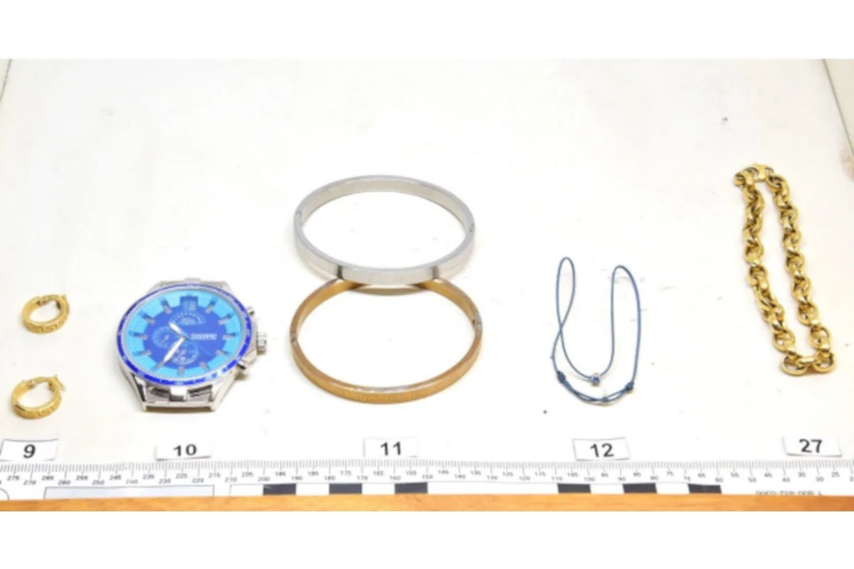 Nahaufnahme einer blauen Uhr und mehreren Schmuckstücken, die auf einem Tisch mit Maßband liegen, möglicherweise aus einer polizeilichen Sicherstellung.