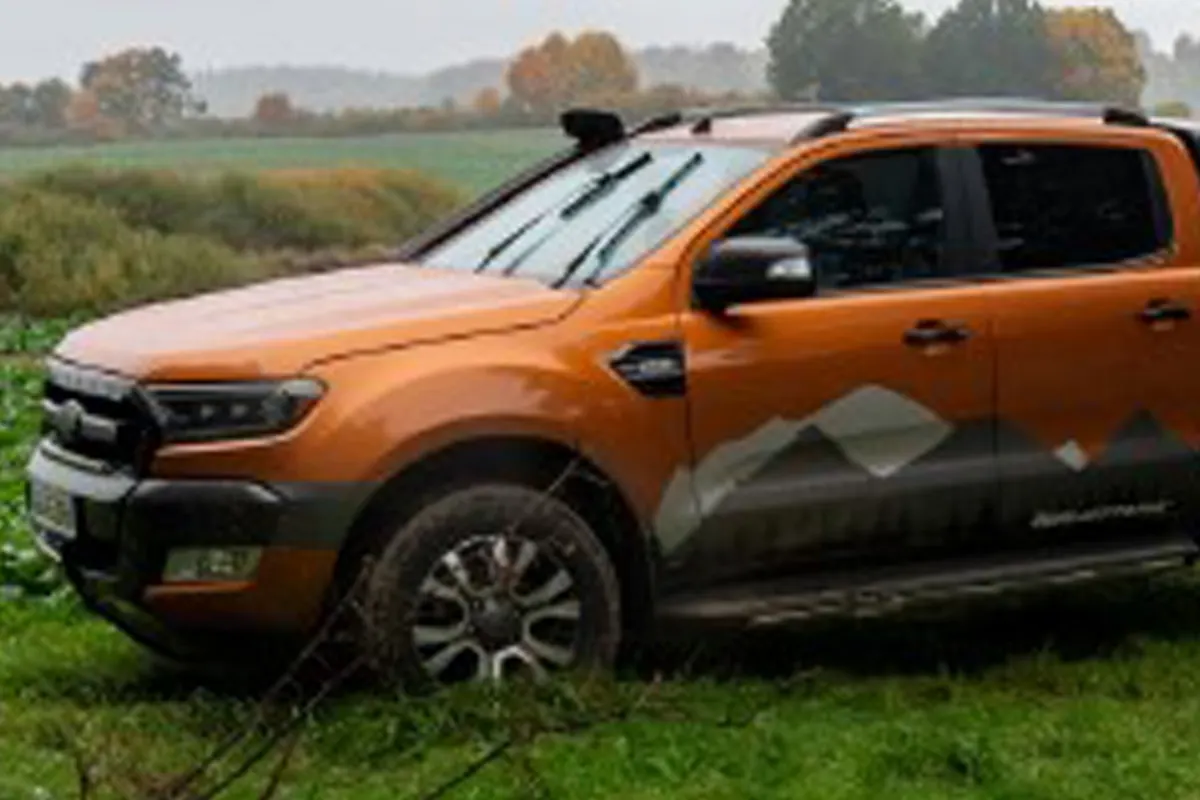 Ein orangefarbener Pickup mit speziellen Markierungen steht auf einer grasbewachsenen Wiese. Bäume sind im Hintergrund sichtbar, was auf eine ländliche Umgebung hindeutet.