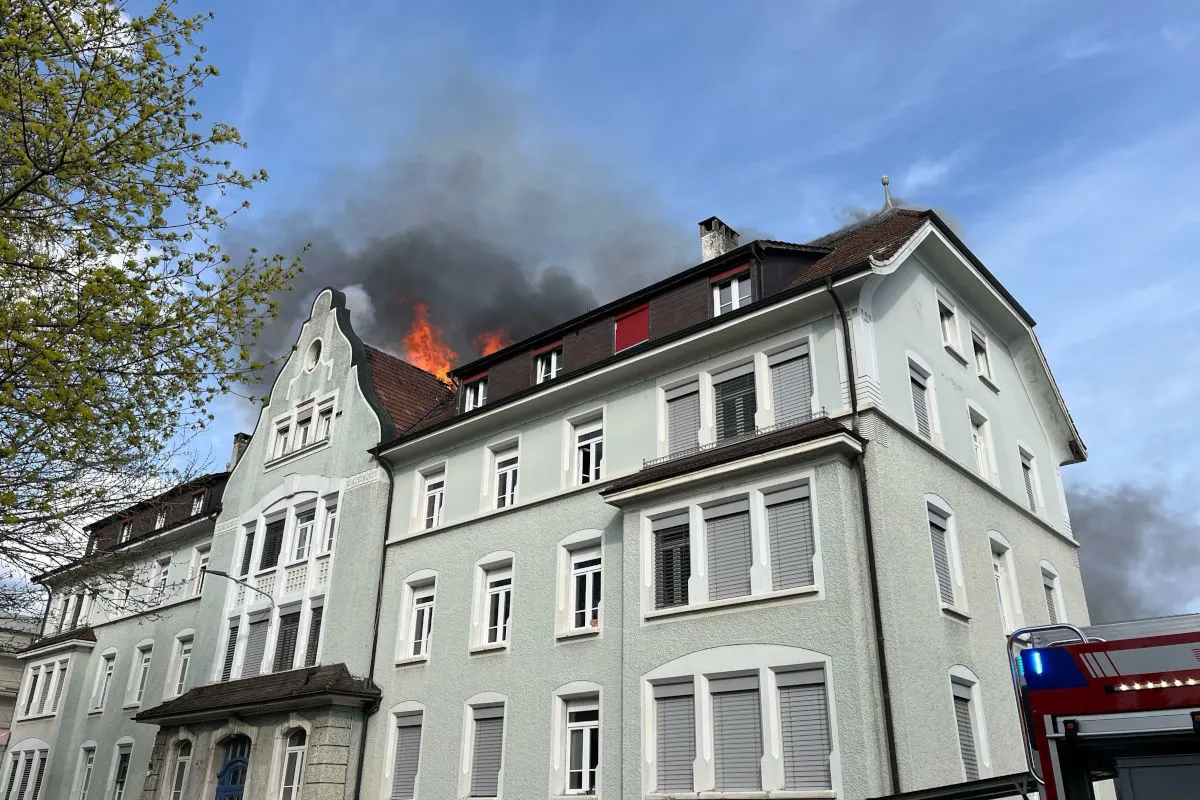 Das Dachgeschoss eines mehrstöckigen Gebäudes brennt stark, mit sichtbaren Flammen und Rauch. Ein Feuerwehrfahrzeug ist am unteren Bildrand erkennbar.