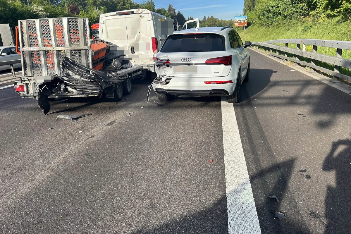 Ein weisser Audi steht beschädigt auf der rechten Fahrspur einer Autobahn. Daneben ist ein beschädigter Transporter mit Anhänger, einige Trümmer liegen auf der Fahrbahn.