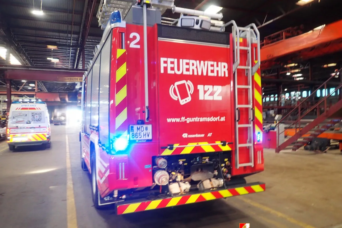 Ein rotes Feuerwehrauto mit der Aufschrift 'FEUERWEHR 122' steht in einer großen Halle. Ein Krankenwagen ist weiter vorne sichtbar.