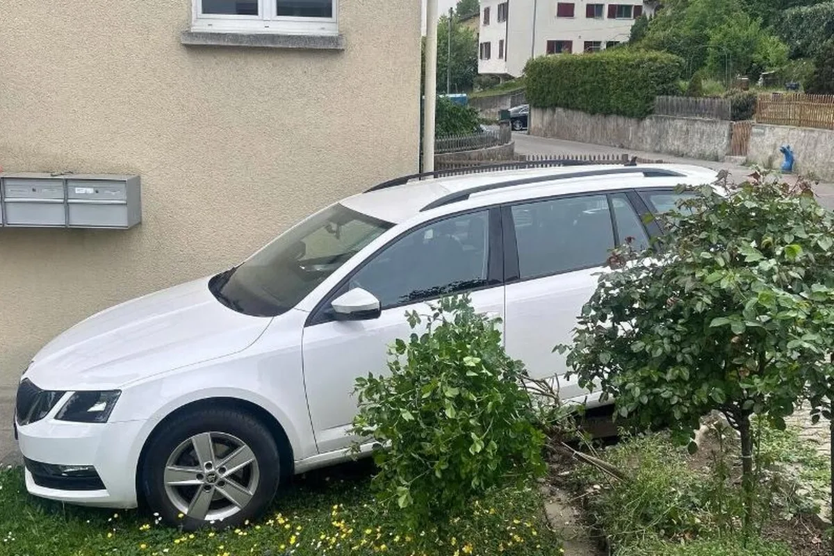Ein weißer Kombi steht auf einer kleinen Grünfläche direkt an einer Hauswand. Im Hintergrund sind weitere Gebäude sichtbar.