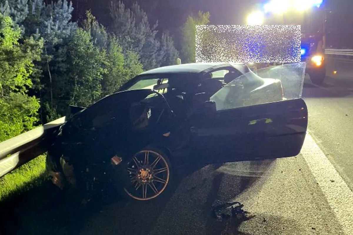 Ein Auto mit Schäden an der Front steht mit geöffneter Tür auf einer nächtlichen Autobahn neben der Leitplanke. Im Hintergrund sind helle Blaulichter sichtbar.