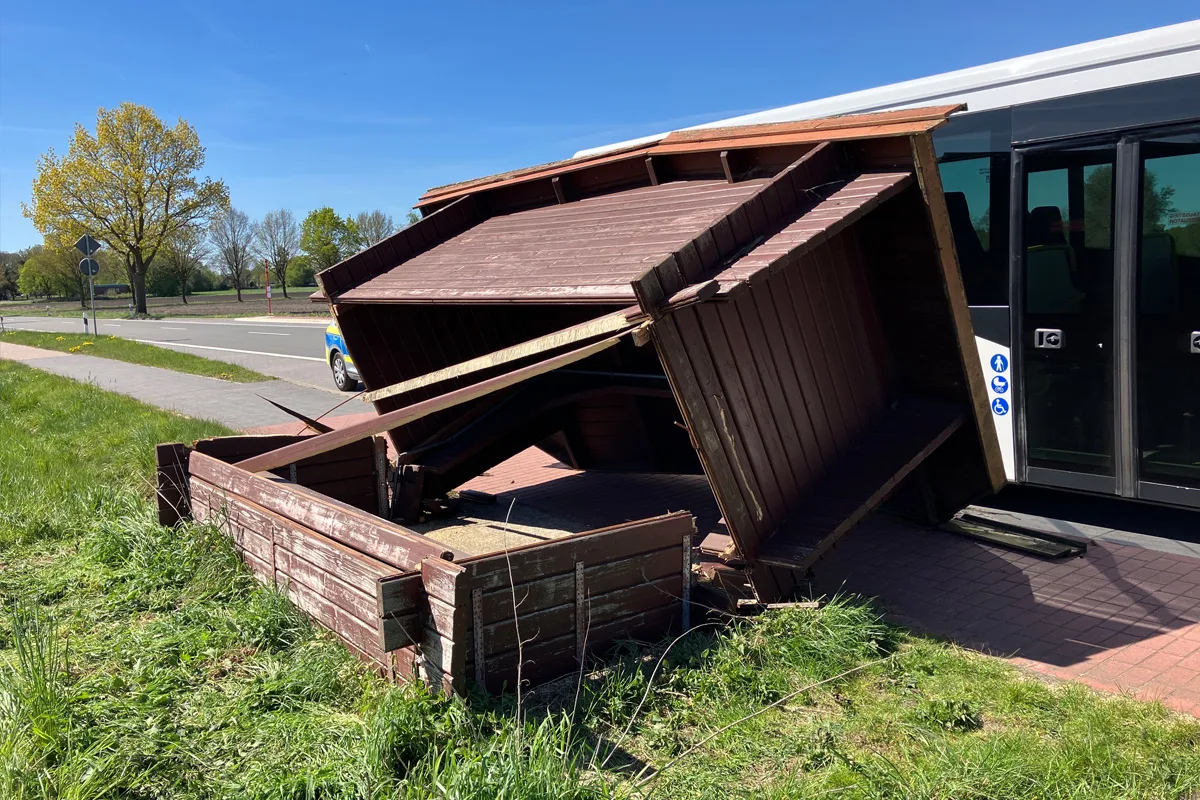 Eine beschädigte Holzhütte liegt umgekippt an einem Bus auf einer Landstraße. Der Himmel ist klar und die Bäume sind grün.