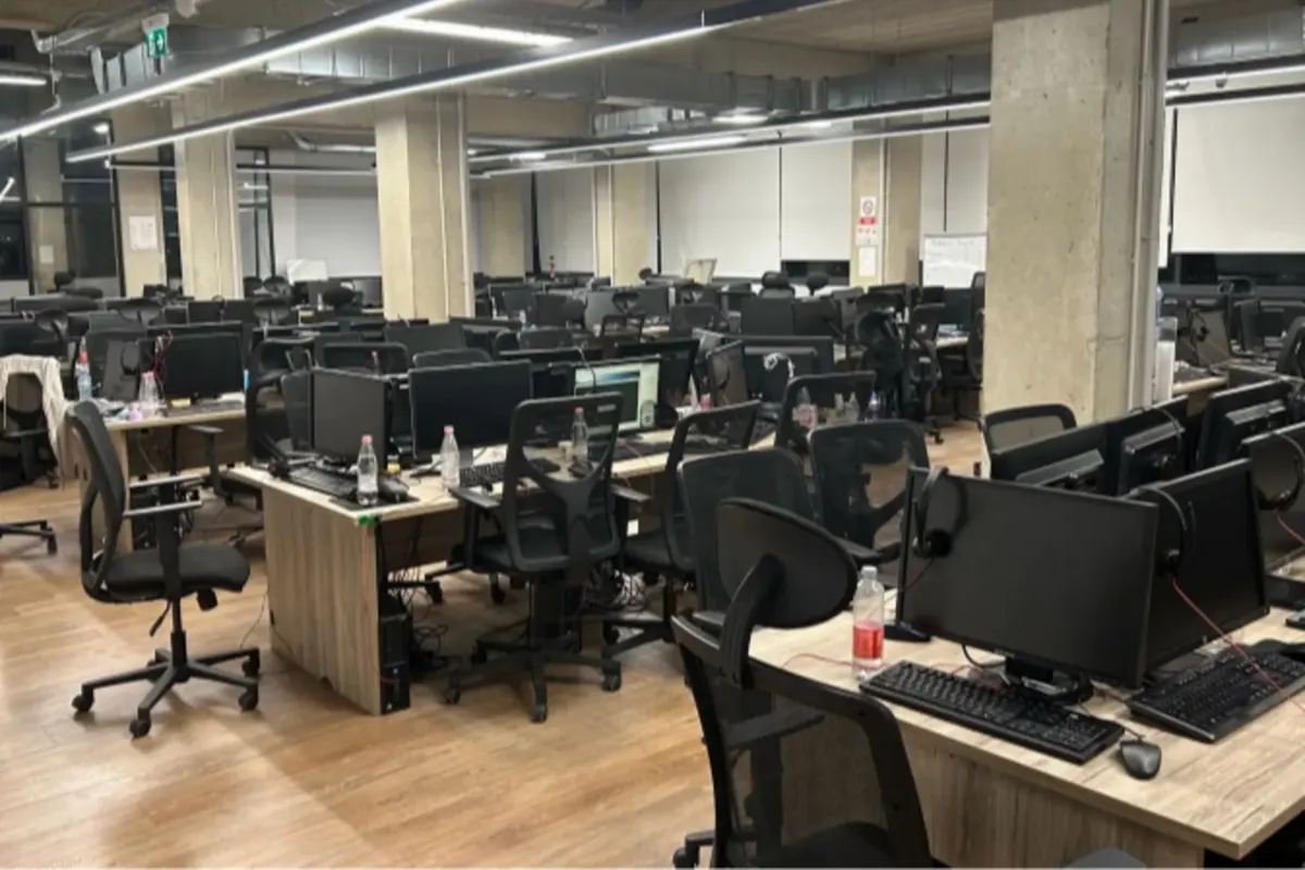 Ein modernes Großraumbüro mit mehreren leeren Schreibtischen, Computern und drehbaren Bürostühlen. Die Beleuchtung ist eingeschaltet und es sind keine Personen anwesend.