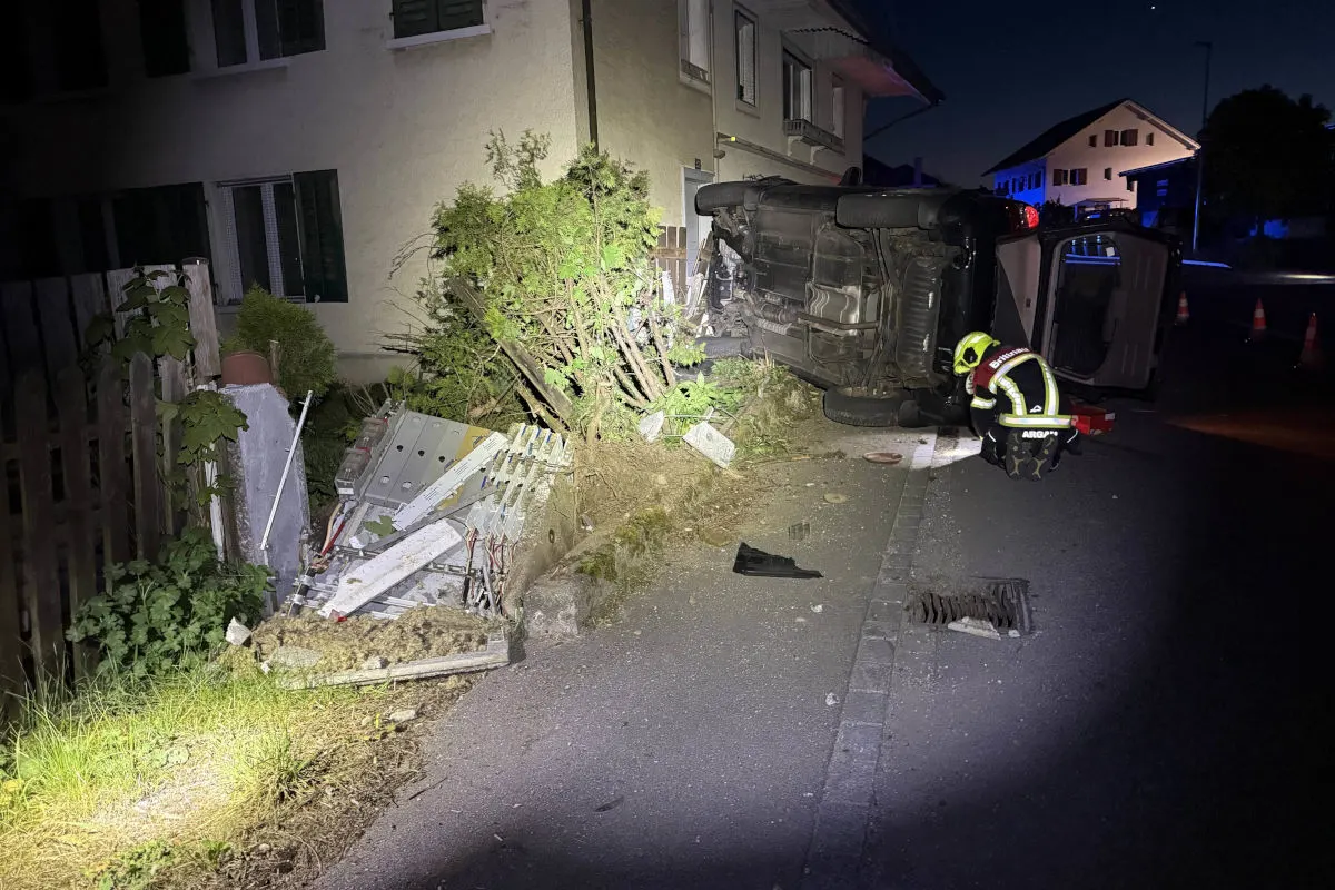 Bei Nacht liegt ein Auto auf der Seite direkt neben einem Haus. Ein Feuerwehrmann kümmert sich um die Situation.