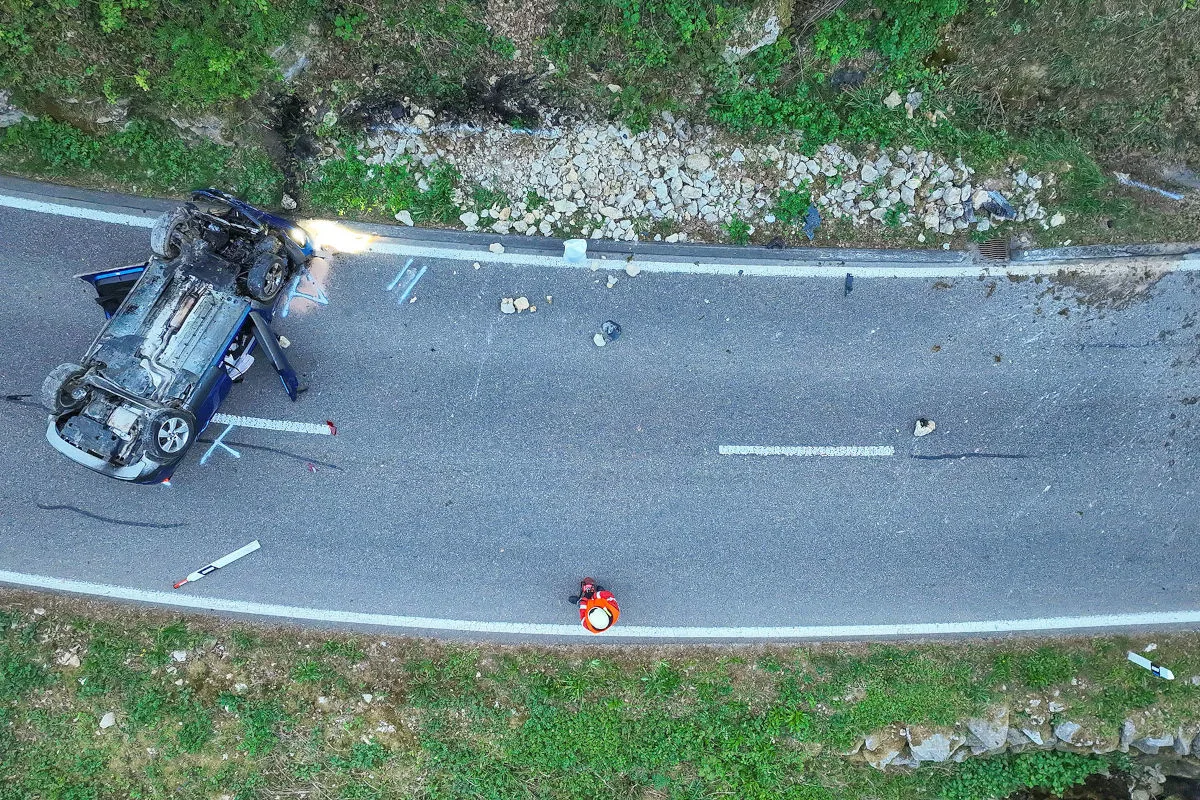 Ein Auto liegt auf dem Dach auf einer zweispurigen Straße, umgeben von Trümmern. Eine Person in Warnweste steht daneben.