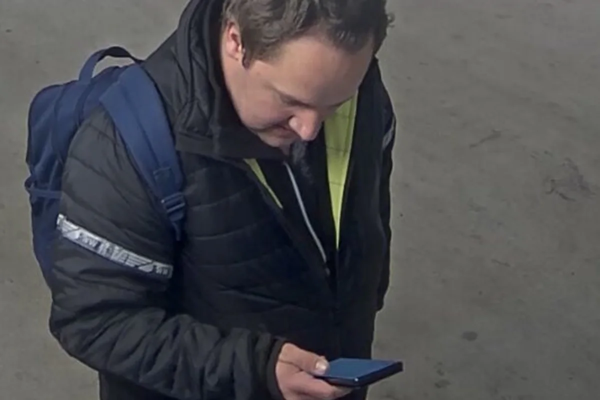 Eine Person steht in einem Innenbereich, trägt eine dunkle Jacke und einen blauen Rucksack, und blickt auf ein Smartphone in der Hand.
