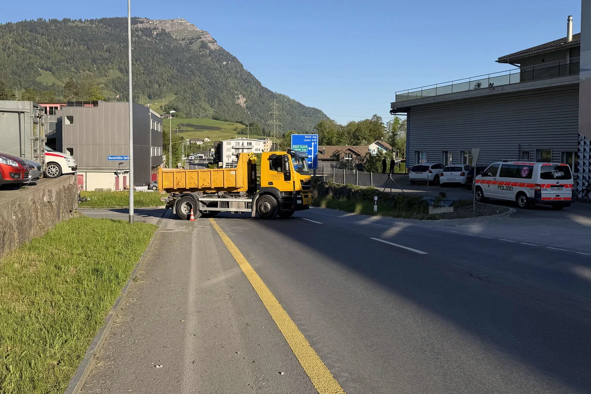 Ein gelber Lastwagen steht auf einer Straße, neben einem parkenden Polizeiauto. Im Hintergrund sind Gebäude und ein Berg zu sehen.