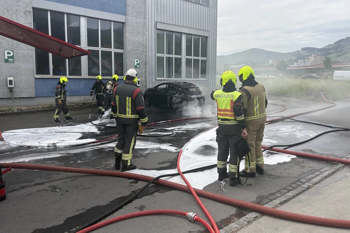 Mehrere Feuerwehrleute, gekleidet in Schutzanzüge, löschen einen brennenden PKW neben einem großen Gebäude. Im Hintergrund sind Berge zu sehen.
