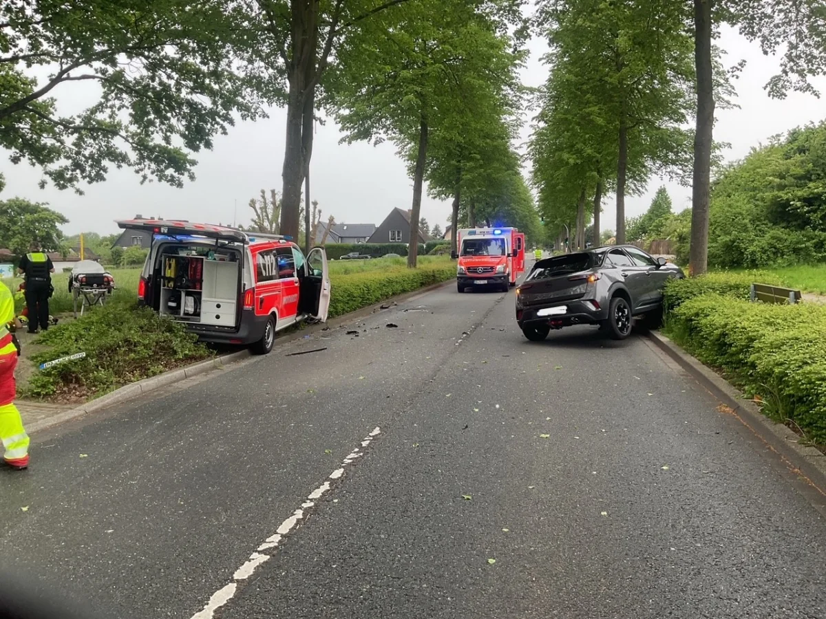 Auf einer Straße sieht man einen Unfall mit einem Rettungswagen links im Graben und einem dunklen Pkw rechts. Personen in Warnwesten stehen daneben.