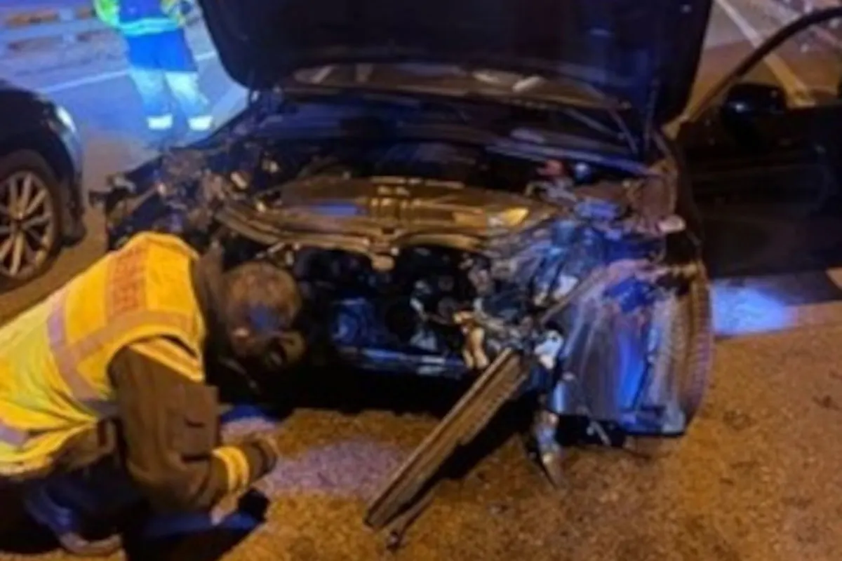 Ein Auto mit geöffneter Motorhaube zeigt deutliche Beschädigungen im Frontbereich. Eine Person in Warnkleidung untersucht den Schaden, während eine weitere Person im Hintergrund steht.