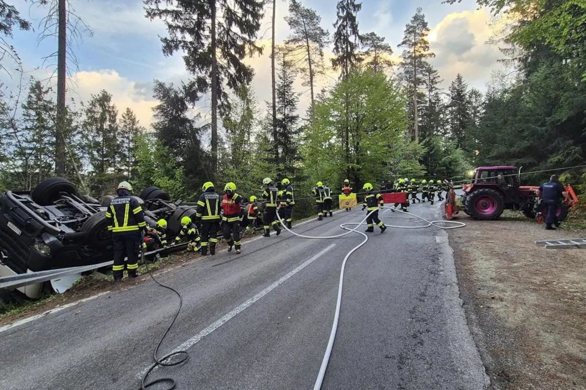 Eine Gruppe von Feuerwehrleuten in leuchtender Schutzkleidung arbeitet an einem umgestürzten Fahrzeug, das auf einer Waldstraße liegt. Im Hintergrund sind Bäume und ein Himmel mit ein paar Wolken zu sehen.