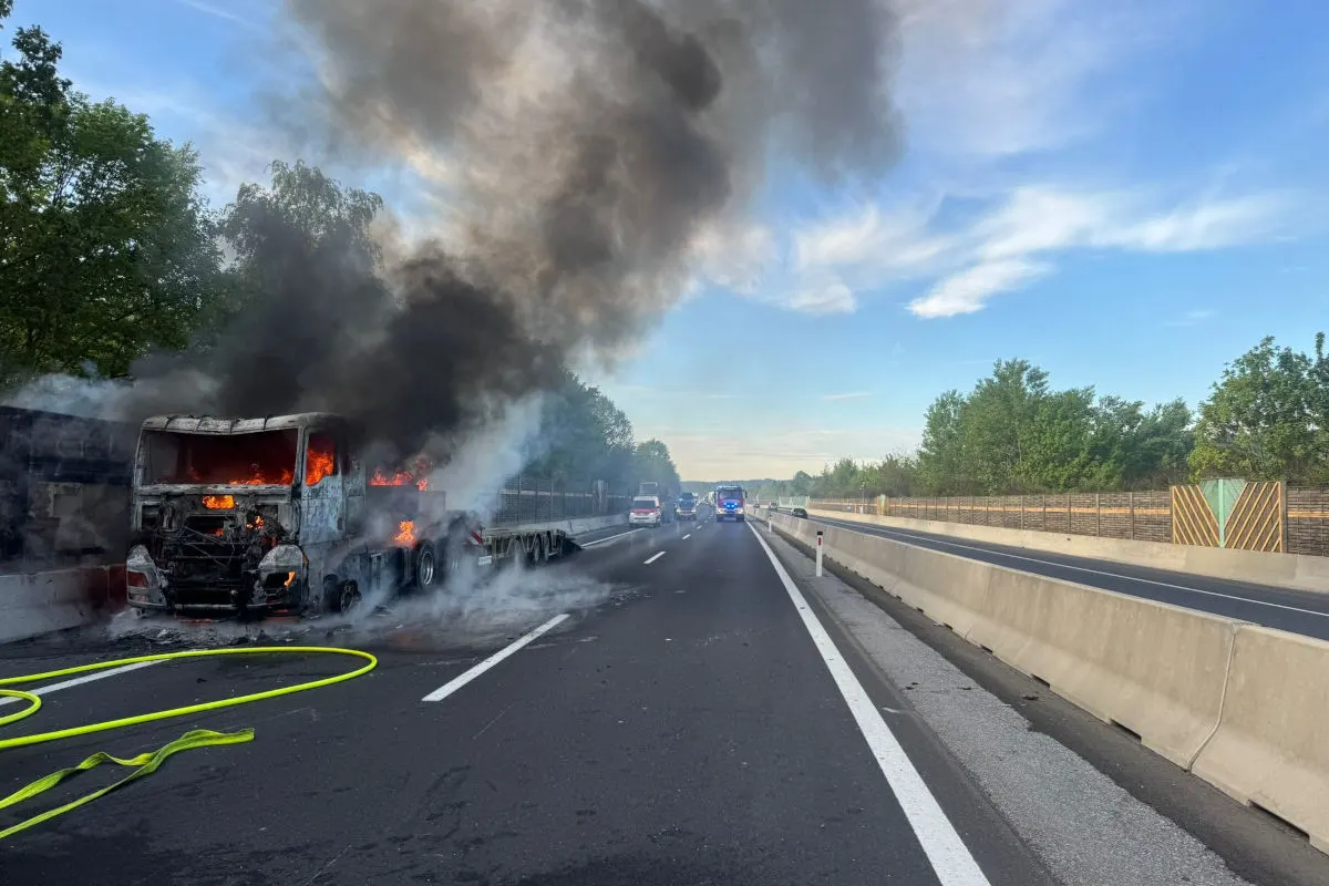 Ein Lastwagen steht in Flammen auf der Autobahn. Dicker schwarzer Rauch steigt in den Himmel. Verkehrsbarrieren und Bäume sind sichtbar.