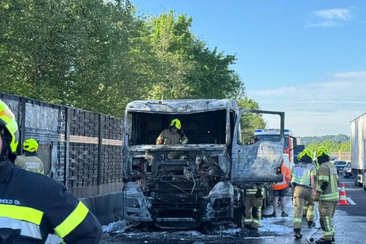 Im Bild ist ein stark beschädigter LKW zu sehen, der von mehreren Feuerwehrleuten betreut wird. Der LKW steht auf einer Autobahn und zeigt schwere Brandschäden.