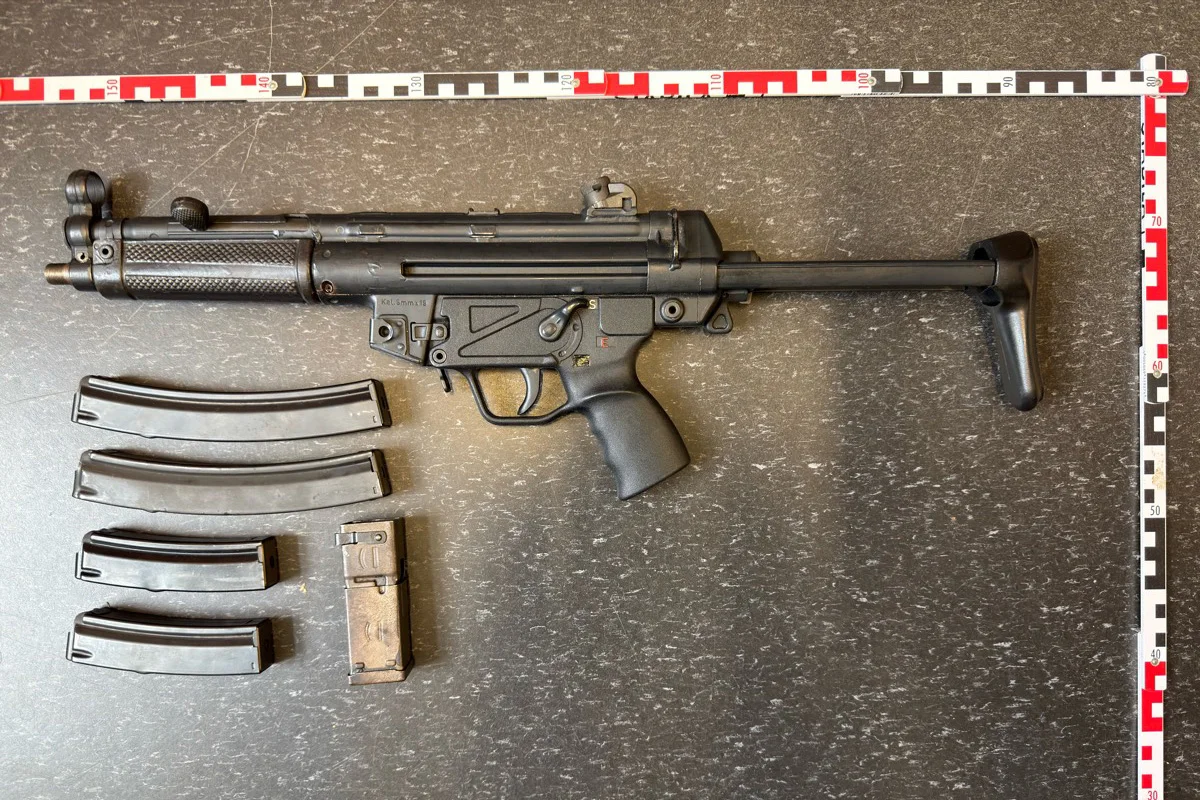 Ein schwarzes Maschinengewehr liegt auf einem dunklen Tisch, daneben mehrere Magazine. Maßstabslineal daneben positioniert.