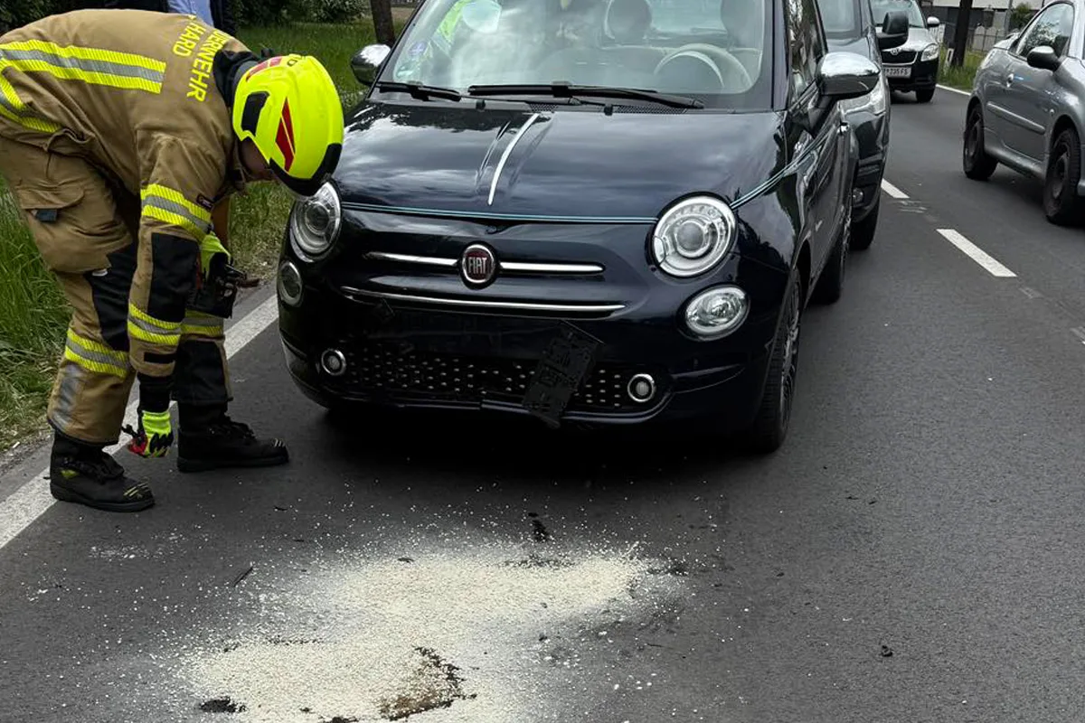 Ein Feuerwehrmann mit gelbem Helm und Uniform steht vor einem schwarzen Auto auf der Straße. Auf dem Boden liegt ein heller, körniger Stoff.