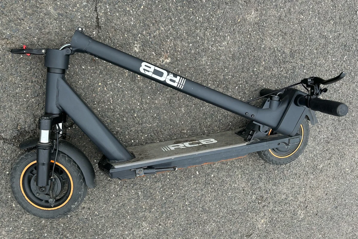 Ein schwarzer E-Scooter liegt quer auf einer Oberfläche aus Asphalt. Der Scooter hat orangefarbene Felgen.