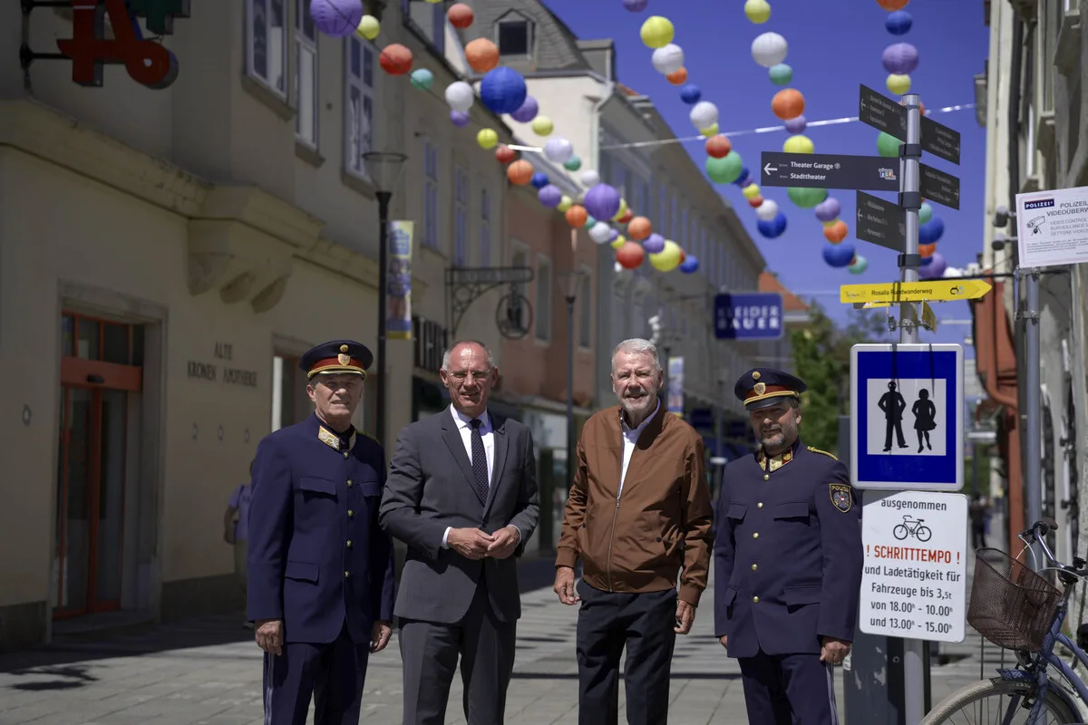 Vier Personen, drei in Uniform und eine in Zivilkleidung, stehen in einer dekorierten Straße mit bunten Laternen über ihnen. Im Hintergrund sind Gebäude und einige Schilder sichtbar.