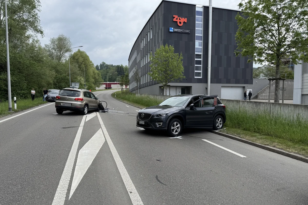 Zwei Autos sind auf einer Straße in einem Verkehrsunfall verwickelt, nahe einem großen Gebäude. Personen stehen am Straßenrand und beobachten die Szene.