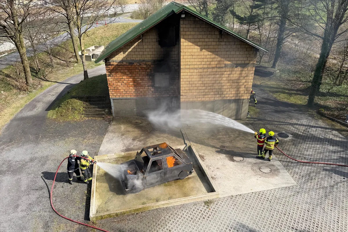 Vier Feuerwehrleute bekämpfen ein Feuer an einem Autowrack vor einem Gebäude. Wasserstrahlen sind sichtbar.