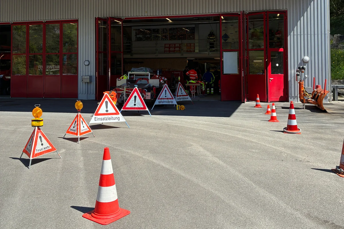 Ein Feuerwehrgebäude mit geöffneter Türen und mehreren Warnschildern sowie Pylonen auf dem Vorplatz, deutet auf einen Einsatz hin. Innen sind Personen in reflektierenden Westen erkennbar.