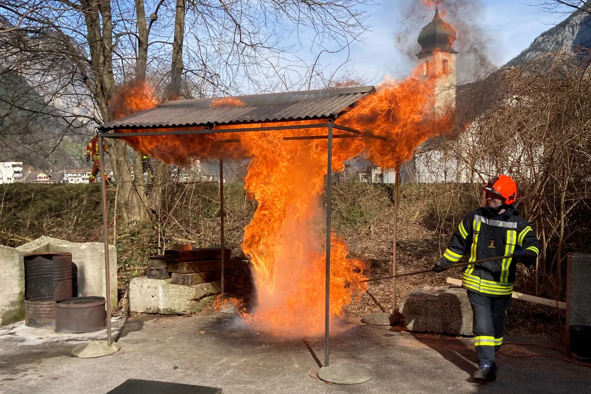 Ein Feuerwehrmann in Schutzkleidung und rotem Helm löscht ein großes Feuer unter einem Blechdach in einer ländlichen Umgebung. Im Hintergrund sind Bäume und ein Kirchturm sichtbar.