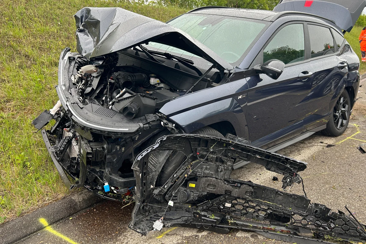 Ein stark beschädigtes Fahrzeug steht am Straßenrand. Die Motorhaube ist geöffnet und Teile der Front sind abgerissen.