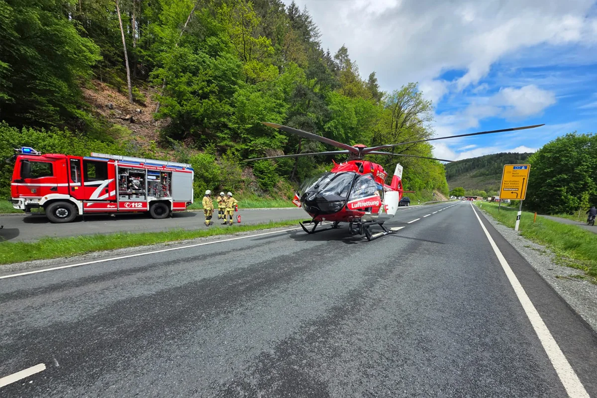 Ein Rettungshubschrauber steht auf einer Landstraße, mit einem Feuerwehrfahrzeug und Einsatzkräften im Hintergrund. Der Hubschrauber ist rot-weiß und trägt die Aufschrift 'Luftrettung'.