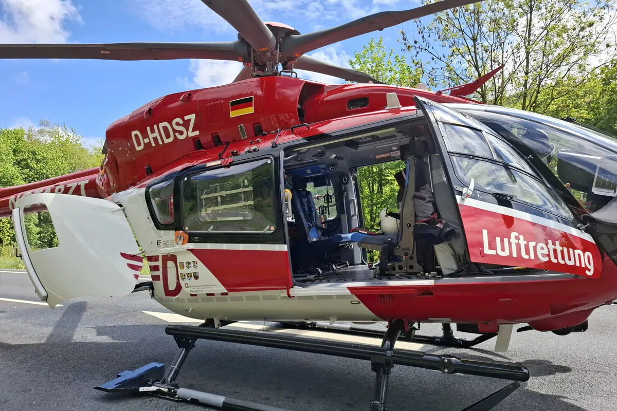 Ein rot-weißer Rettungshubschrauber steht am Straßenrand. Die seitliche Tür ist geöffnet und zeigt das Innere des Hubschraubers. Es ist tagsüber mit klarem Himmel und grünem Laub im Hintergrund.