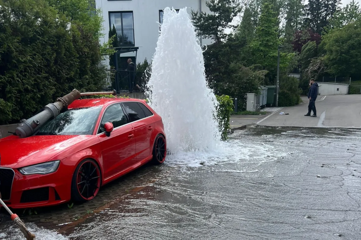 Ein rotes Auto steht schräg neben einem Hydranten, der Wasser auf die Straße spritzt. Im Hintergrund sind Bäume und ein Wohnhaus zu sehen, während ein Mann am Straßenrand steht.