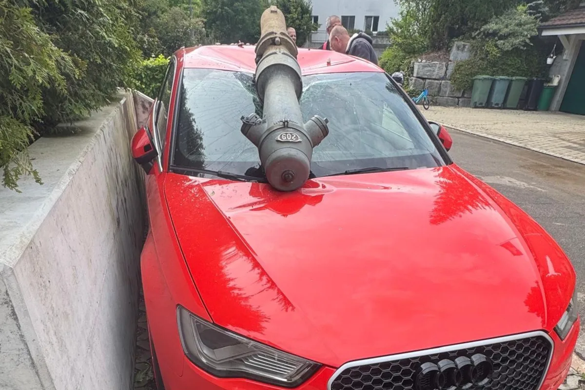 Ein roter Audi steht in einer Wohngegend mit einem abgebrochenen Hydranten, der die Windschutzscheibe durchschlägt. Personen stehen in der Nähe des beschädigten Fahrzeugs.