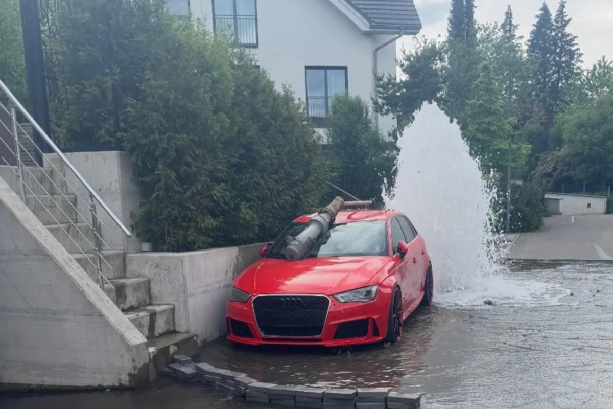 Ein rotes Auto ist neben einem geplatzten Hydranten zu sehen, aus dem eine große Wasserfontäne strömt. Das Auto steht nahe einer Treppe, umgeben von hohen Büschen.