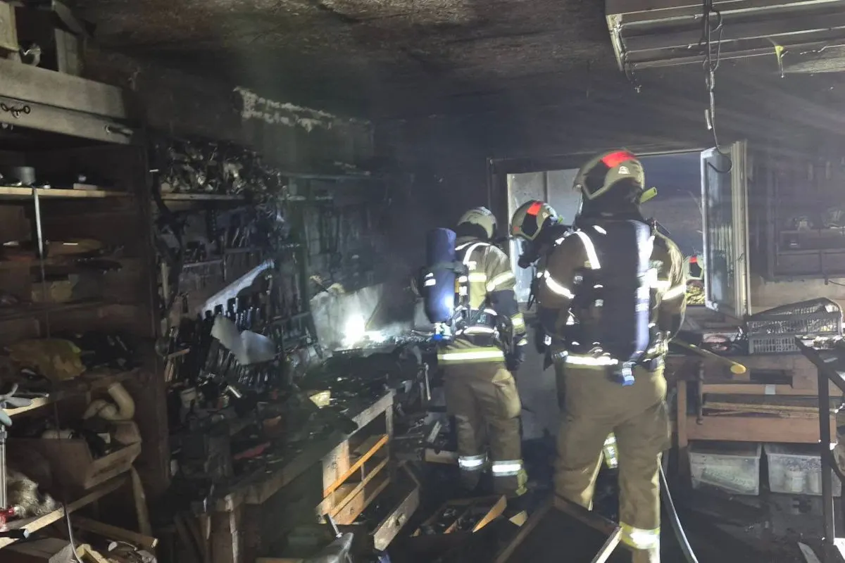 Mehrere Feuerwehrleute in Schutzkleidung agieren in einem rauchgefüllten Raum, offenbar bei einem Einsatz. Der Raum ist mit verschiedenen Werkzeugen gefüllt und zeigt Spuren von Ruß und Brand.