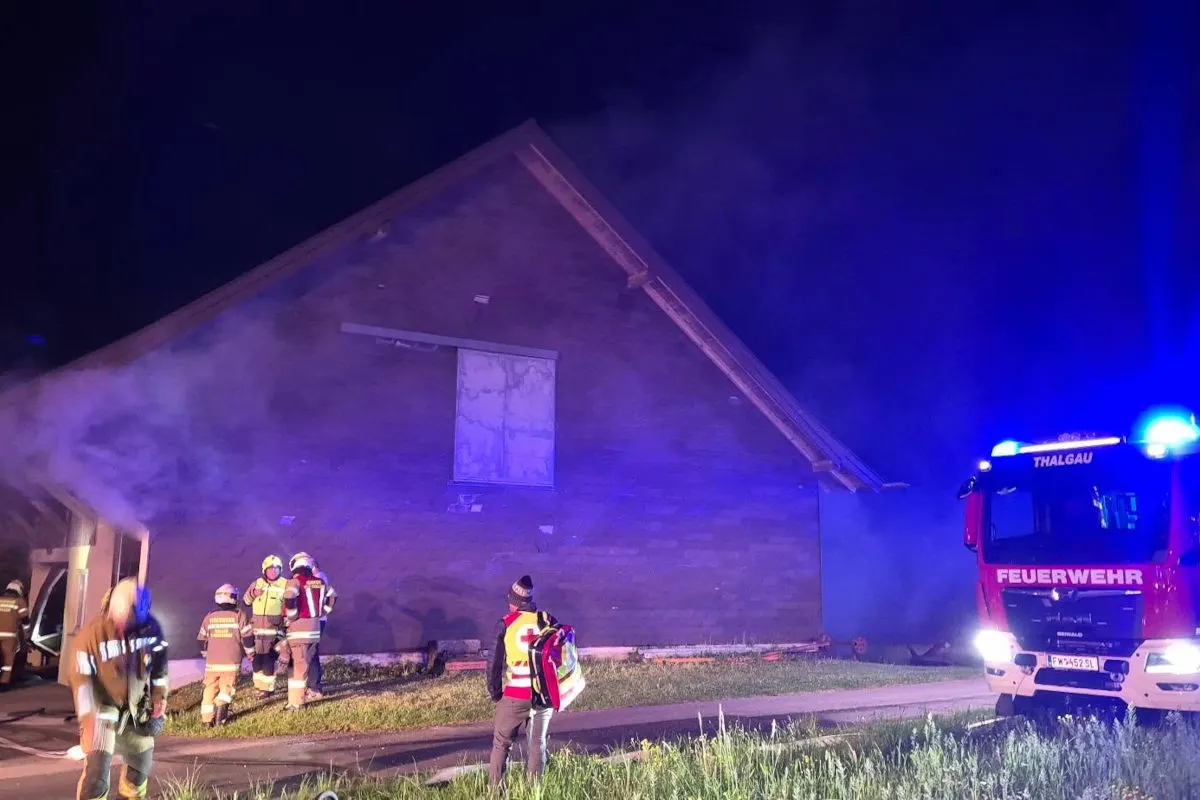 Mehrere Feuerwehrleute stehen vor einem Gebäude, aus dem Rauch austritt. Rechts ist ein rotes Feuerwehrfahrzeug mit eingeschaltetem Blaulicht sichtbar.