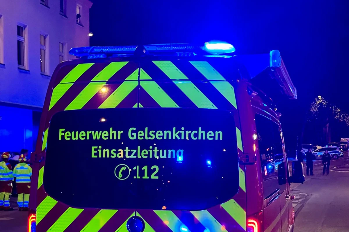 Ein Feuerwehrauto der Einsatzleitung von Gelsenkirchen, leuchtend im Dunkeln mit reflektierenden Streifen, steht auf einer nächtlichen Straße. Im Hintergrund sind weitere Fahrzeuge und Menschen zu sehen.