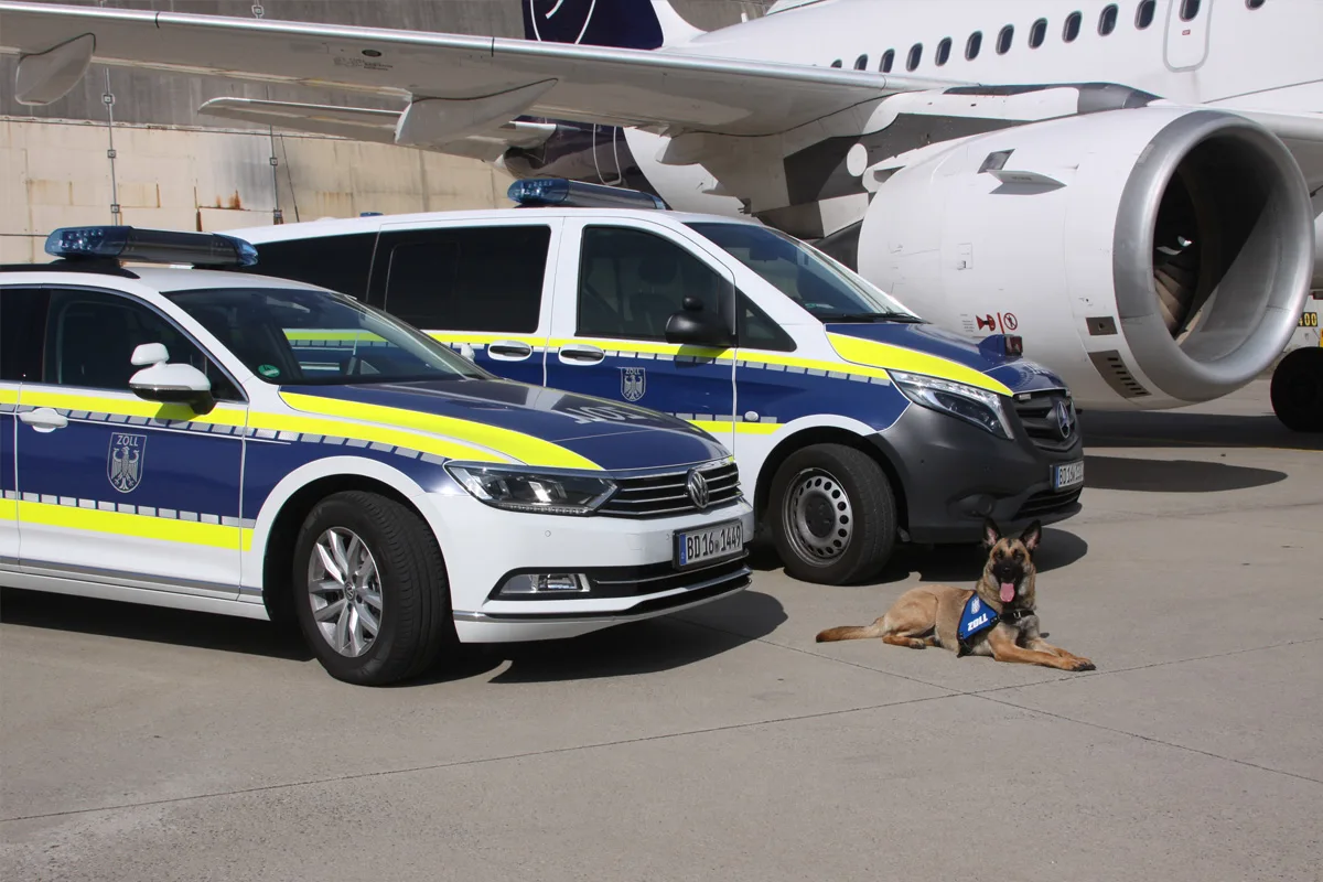 Zwei weiß-blaue Zollfahrzeuge stehen mit eingeschaltetem Blaulicht vor einem Flugzeug; ein Hund mit der Aufschrift 'Zoll' sitzt im Vordergrund auf dem Boden.