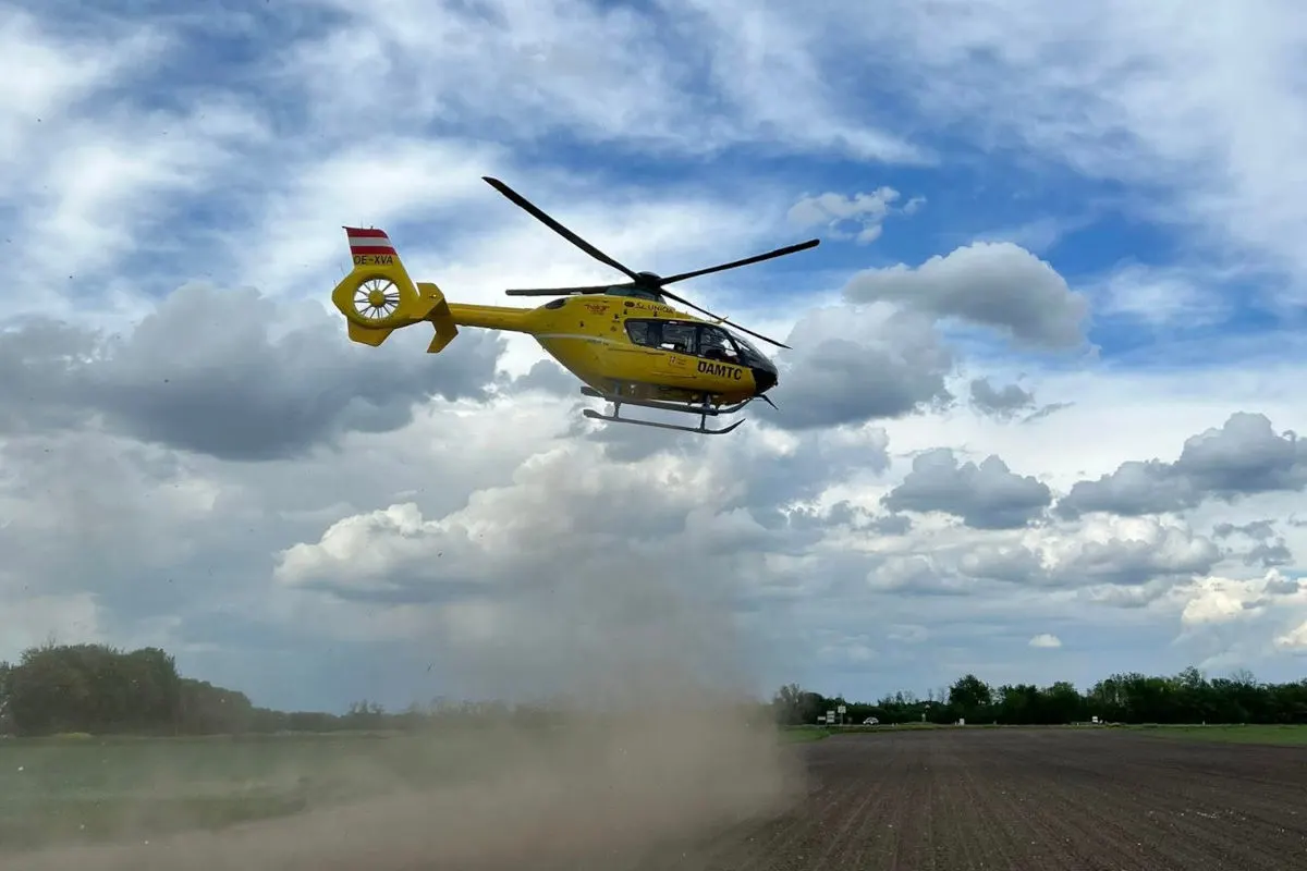 Ein gelber Rettungshubschrauber fliegt über ein teils staubiges, flaches Feld. Graue Wolken sind am Himmel zu sehen.