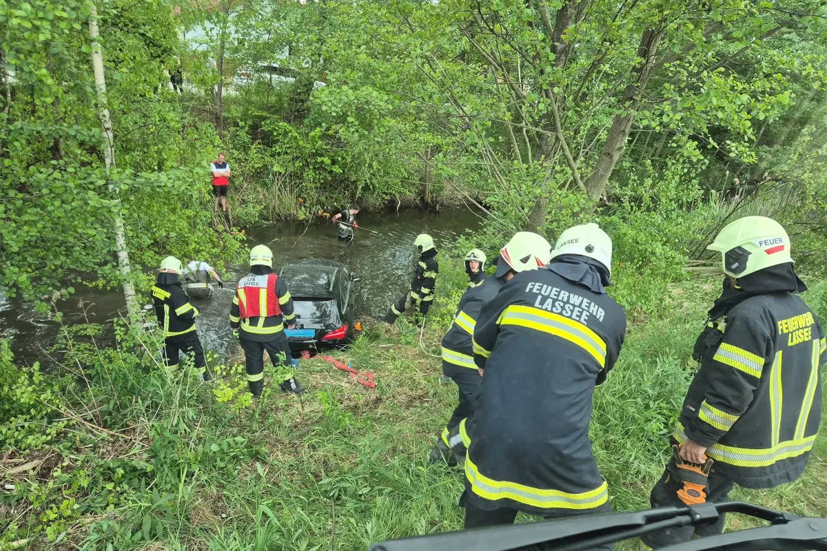 Mehrere Feuerwehrleute stehen am Ufer eines Baches und arbeiten daran, ein Auto aus dem Wasser zu bergen. Das Gelände ist bewaldet und grün.