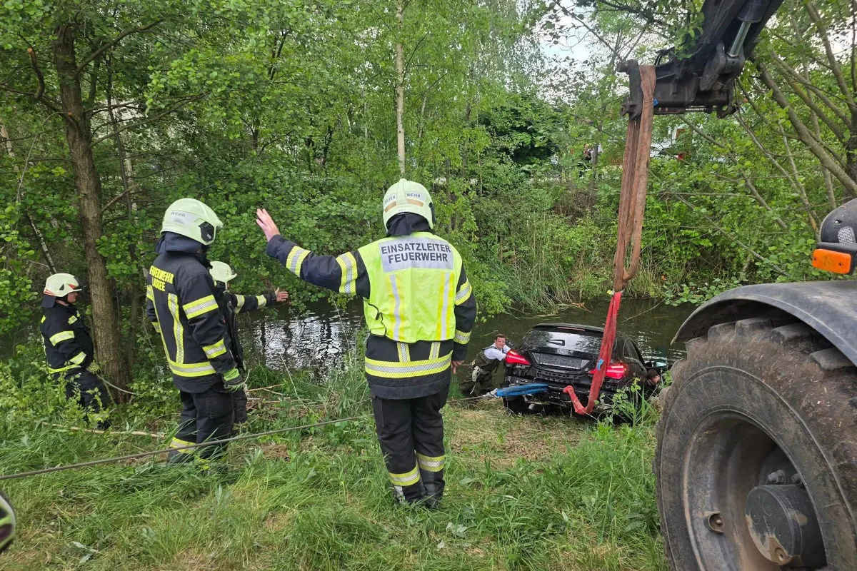 Ein schwarzes Auto wird mit Hilfe eines Krans aus einem Fluss geborgen. Mehrere Feuerwehrleute sind am Ufer im Einsatz.