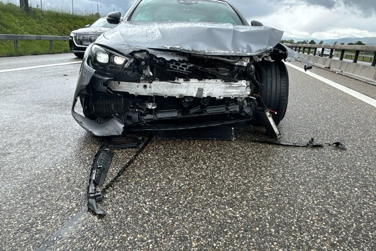 Ein graues Auto mit stark beschädigter Vorderseite steht auf einer nassen Autobahn. Im Hintergrund ist ein weiteres Fahrzeug zu sehen.