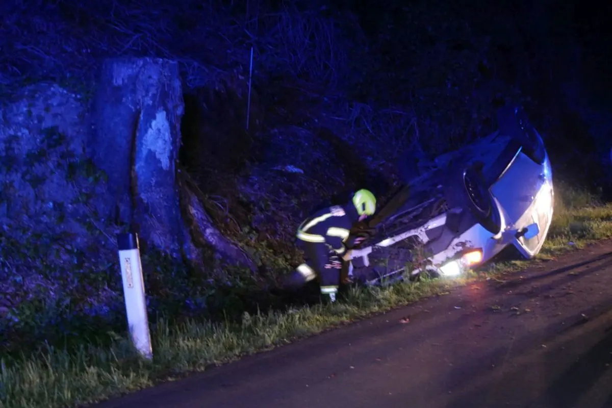Ein Feuerwehrmann mit gelbem Helm untersucht ein Auto, das auf der Seite an einer Böschung liegt. Das Bild ist nachts aufgenommen.